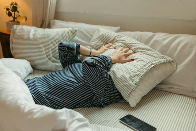 Someone lying face down on a bed with a pillow pulled over their head, phone abandoned on the sheets beside them.